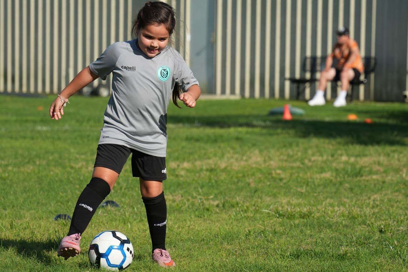 A young NEXO player in a grey club shirt smiles while dribbling a soccer ball on grass
